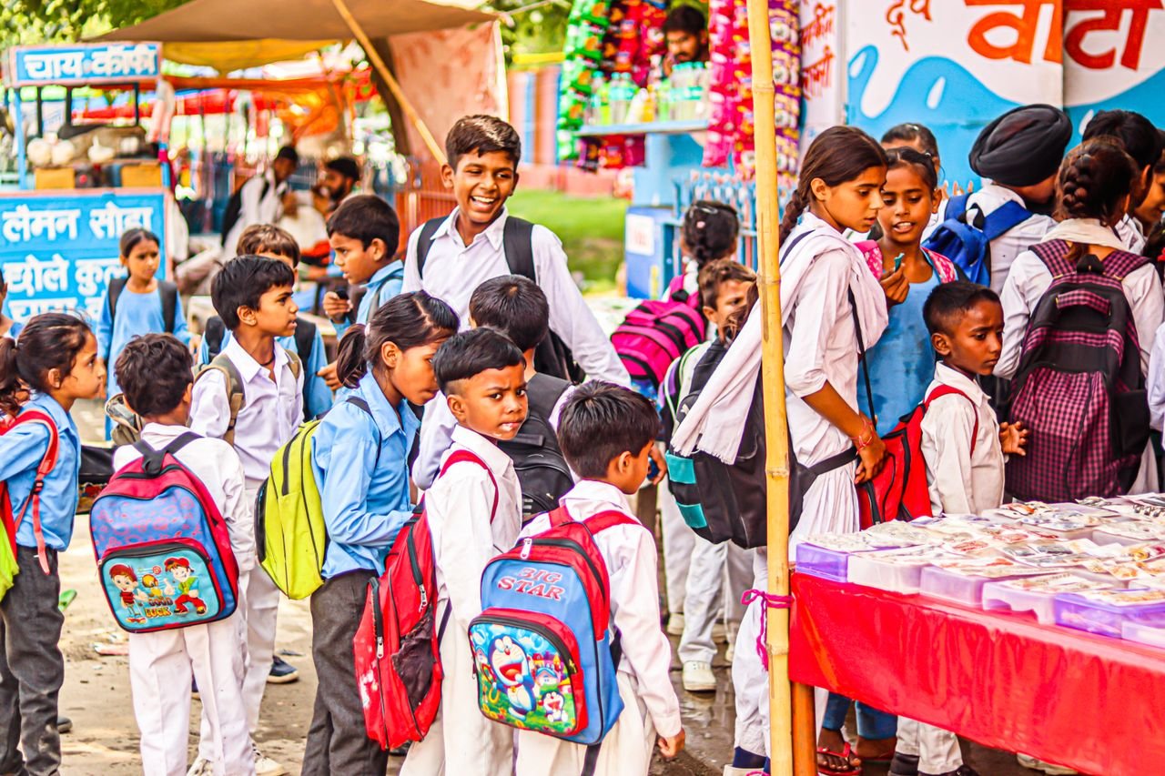 Group of children in uniforms with backpacks queuing at a market stall outdoors.