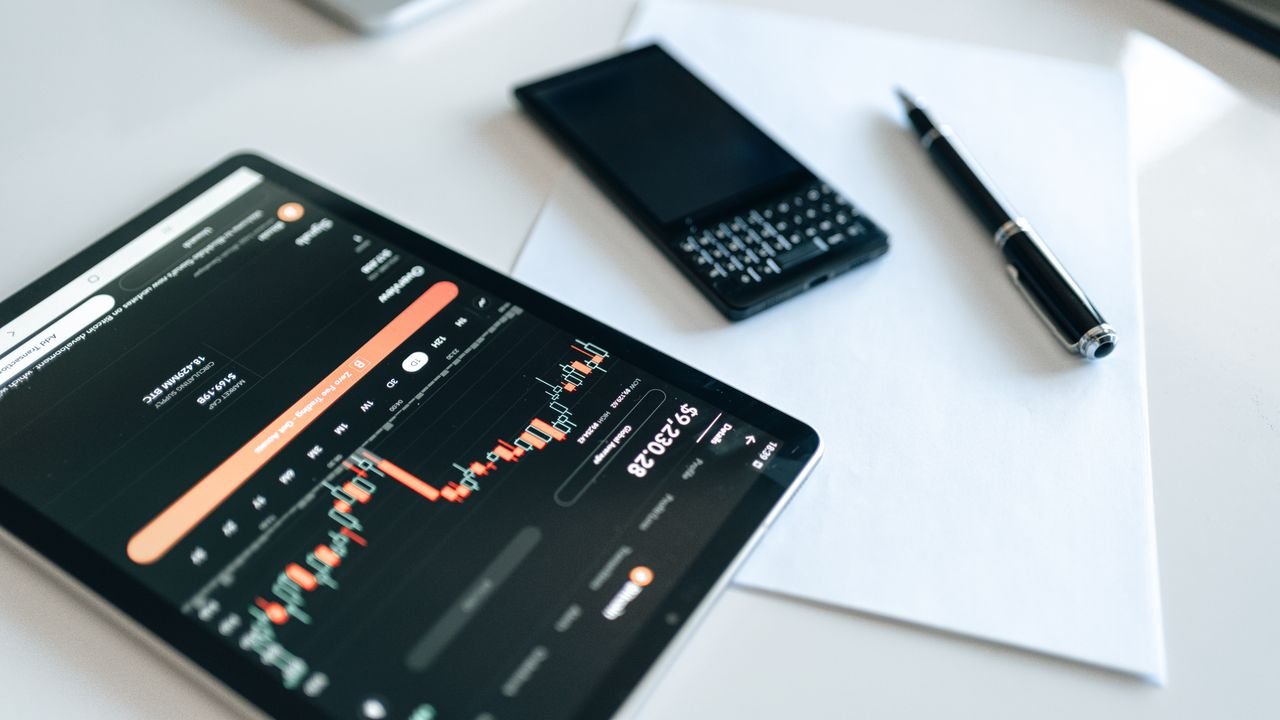 Close-up of a tablet displaying financial charts with a phone and pen on a white surface.